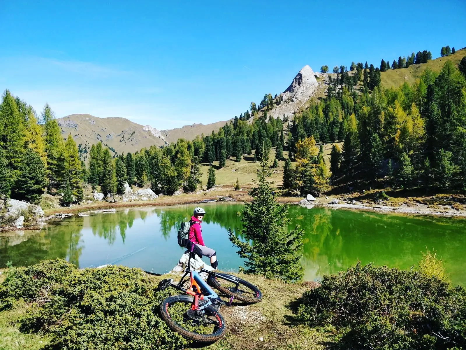 Ciclista con e-MTB accanto a un lago alpino verde, con larici e una cima dietro