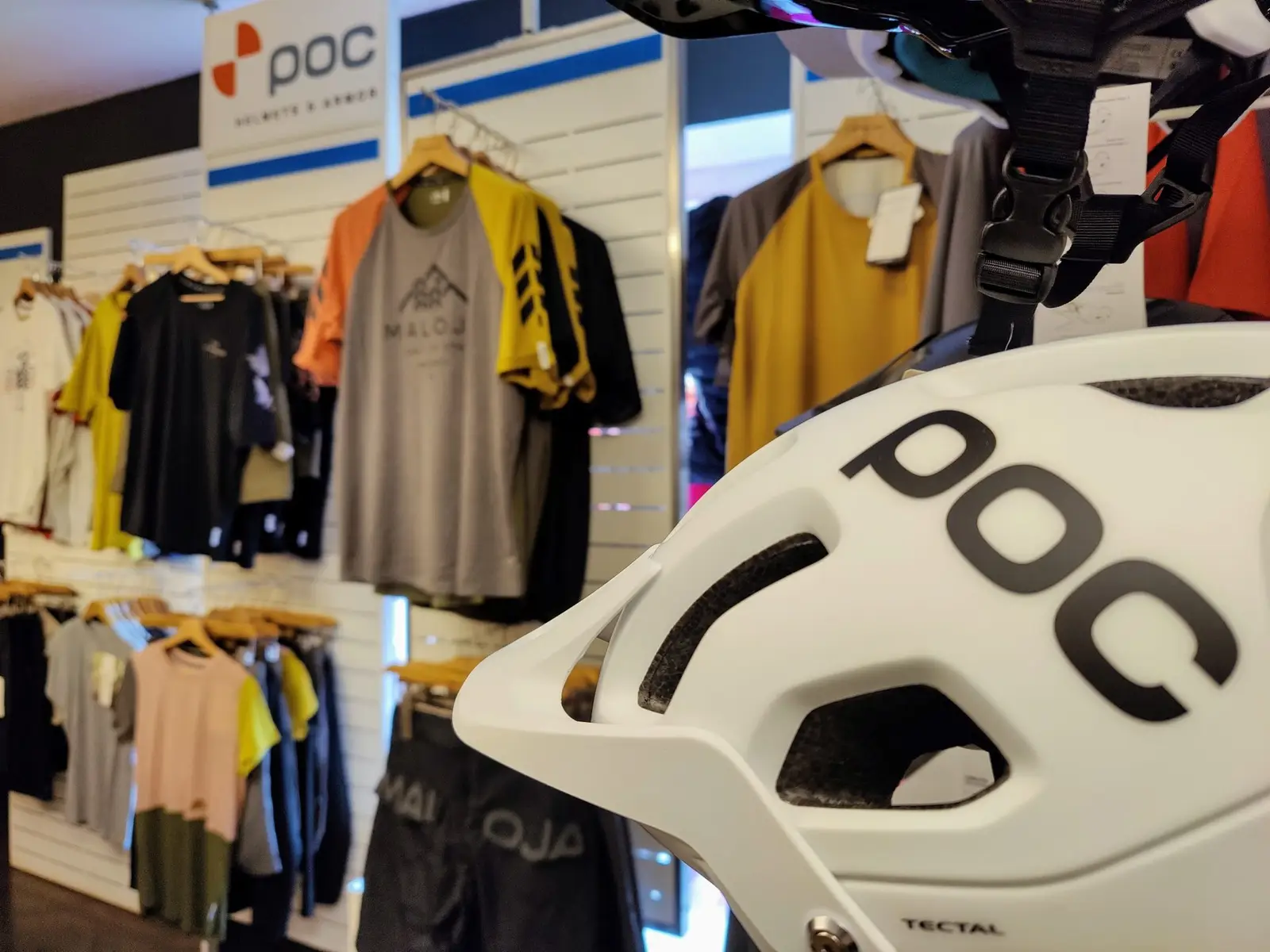 Interno del negozio: casco da bici POC bianco in primo piano, magliette Maloja alla parete