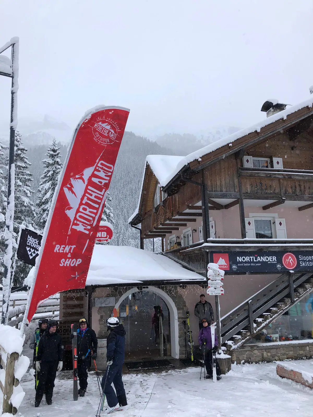 Eingang des Geschäfts bei Schneefall mit roter Fahne „Northland Rent & Shop“ und Skifahrern davor