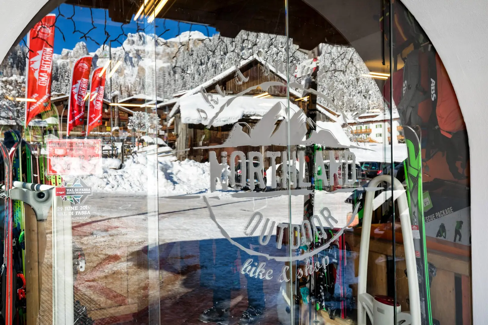 Blick durch die Glastür des Geschäfts mit dem Aufkleber „Northland Outdoor bike & shop“, draußen das verschneite Canazei und rote Fahnen