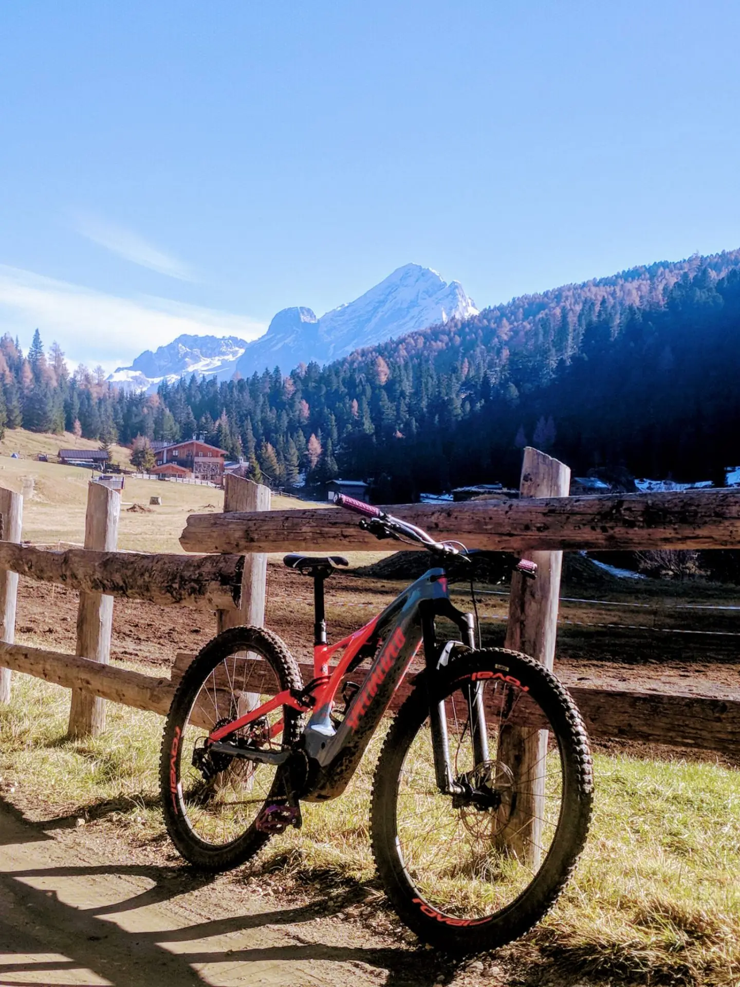 E-MTB rossa e nera appoggiata a una staccionata di legno, larici autunnali e una cima innevata