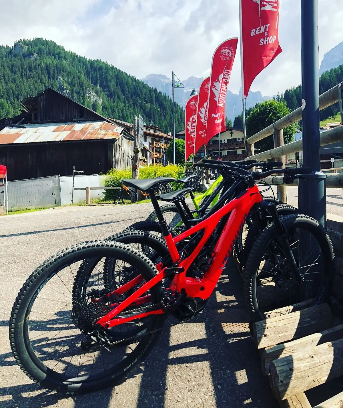 E-MTB rosse parcheggiate davanti al negozio sotto le bandiere rosse Northland, con le case di Canazei dietro