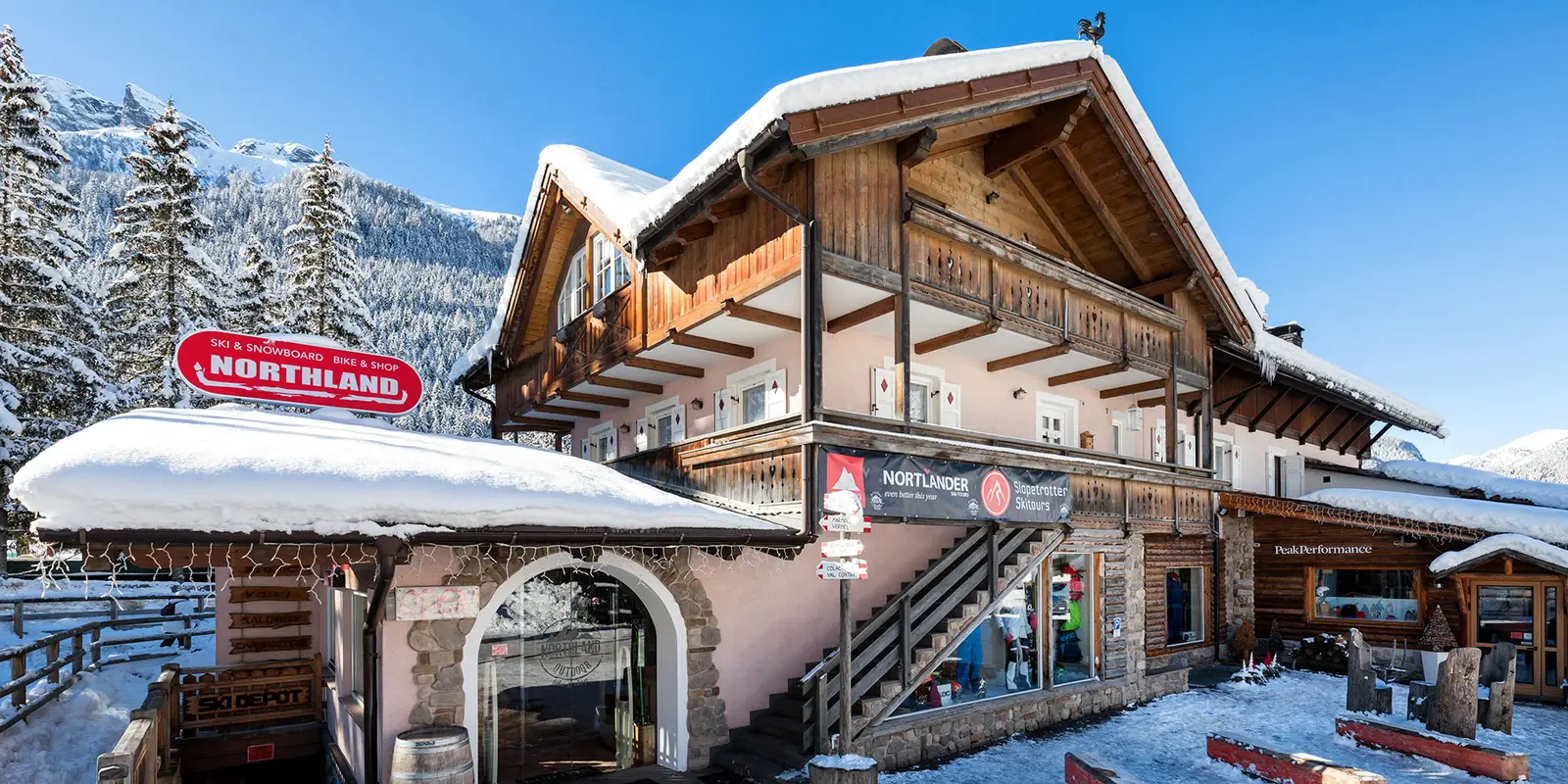 Außenansicht des Northland-Geschäfts im Winter: Holzchalet mit verschneitem Dach, rotes „Northland“-Schild, blauer Himmel