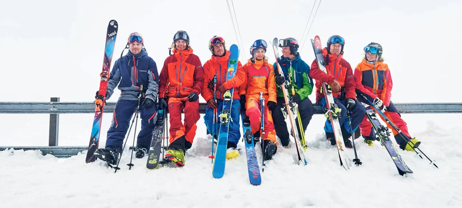 Sieben Skifahrer in bunter Skibekleidung sitzen im Schnee auf einer Leitplanke und halten ihre Ski